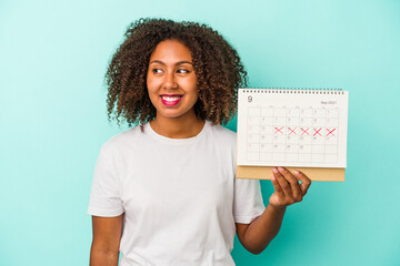 Young african american woman holding a calendar isolated on blue background looks aside smiling,...