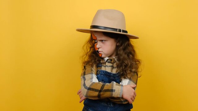 Close up portrait of angry sad little girl kid with halloween makeup, wears brown hat and shirt, crossed arms looking to side, posing isolated over yellow studio background wall with copy space