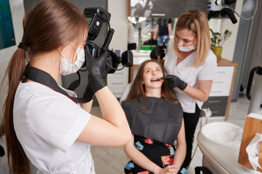 Dentist examining woman teeth while assistant in medical mask taking photo with professional camera. Young female doctor in sterile gloves photographing patient and dentist in dental office.