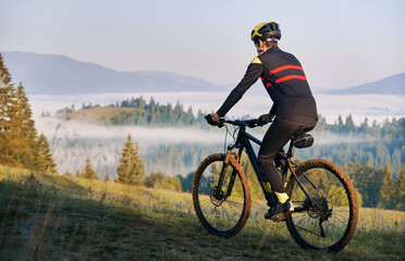 Back view of male cyclist in cycling suit riding bike on road. Man bicyclist wearing safety helmet while enjoying bicycle ride in mountains in the morning. Concept of sport, biking and active leisure.