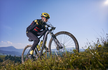 Young man cycling uphill on sunny day with blue sky on background. Male bicyclist in cycling suit climbing uphill on mountain bike. Concept of sport, mountain biking and active leisure.