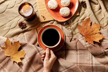 people, season and leisure concept - cup of coffee, autumn leaves, meringues and candle on warm blankets at home