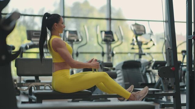 Hispanic woman does exercises on the rowing machine. A girl sits behind a simulator is called a rowing machine. sitting pulls the weight of large Windows and treadmills of the gym