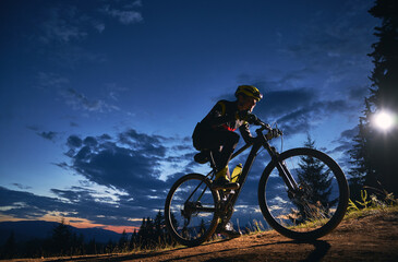 Fototapeta na wymiar Young man cycling bicycle under beautiful night sky. Male bicyclist in safety helmet riding on hillside road under blue cloudy sky at night. Concept of sport, biking and active leisure.