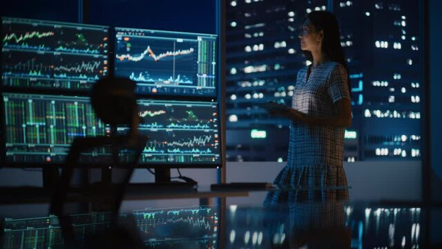 Financial Analyst Using Tablet Computer, Standing Next to Multi-Monitor Workstation with Real-Time Stocks, Commodities and Exchange Market Charts. Businesswoman Working in Investment Bank City Office.