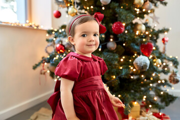 winter holidays and childhood concept - lovely baby girl in red dress over christmas tree at home