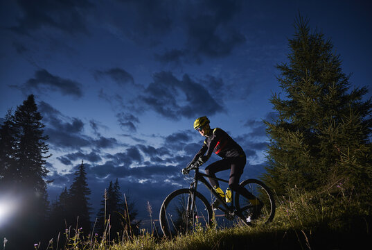 Man cyclist riding bicycle downhill with beautiful blue evening sky on background. Male bicyclist in sports cycling suit cycling down grassy hill at night. Concept of sport, biking and active leisure.