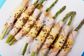 Baked asparagus wrapped with ham, cheese, flaxseeds and soy sauce on a purple, yellow and blue checkered tablecloth background.