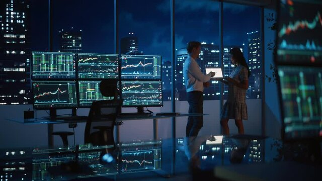 Financial Analyst Talking to Investment Banker in Late Evening at Work. Chatting About Real-Time Stock Chart Data on Tablet Computer. Businesspeople Have a Meeting in Broker Agency City Office.