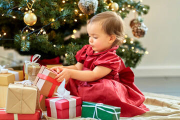 christmas, holidays and childhood concept - happy baby girl opening gifts at home