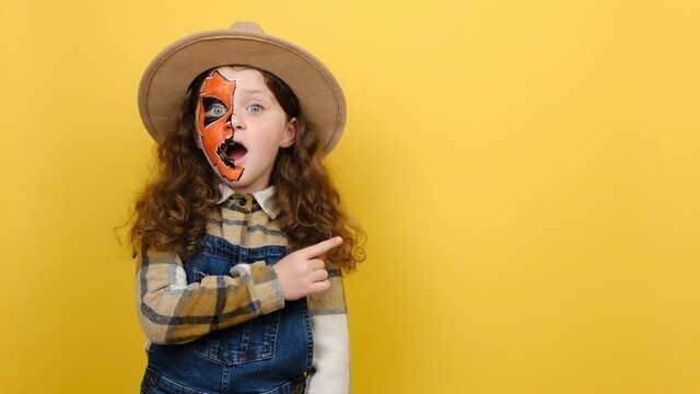 Promoter shocked little girl kid with Halloween makeup mask wears hat and shirt, point finger aside on workspace copy space mockup promo area, posing isolated on yellow color background in studio