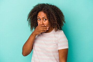 Young african american woman with curly hair isolated on blue background covering mouth with hands looking worried.