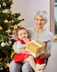 Obraz na płótnie Canvas christmas, holidays and family concept - happy grandmother and baby granddaughter with gift at home