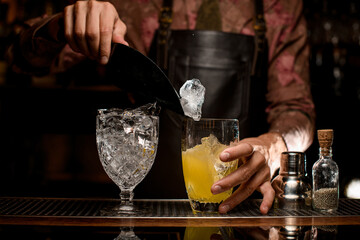Crystal goblet with ice cubes and glass with drink on bar counter. Hand of bartender pours ice into glass using scoop.