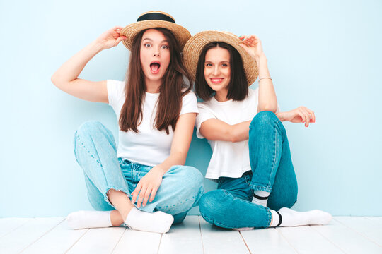 Two young beautiful smiling hipster female in trendy summer white t-shirt and jeans clothes.Sexy carefree women posing near light blue wall in studio.Positive models in hats. Happy and cheerful