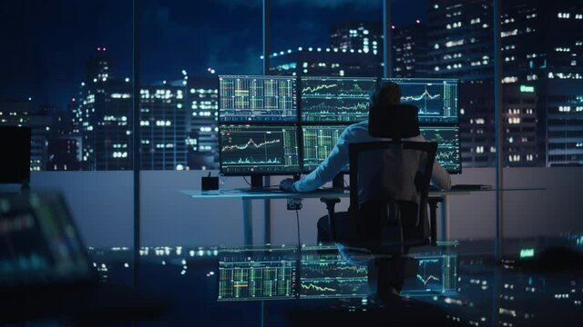 Financial Analyst Working on a Computer with Multi-Monitor Workstation with Real-Time Stocks, Commodities and Exchange Market Charts. Businessman Works in Investment Bank Downtown Office at Night.