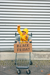 Girl in yellow jacket with arms up in shopping cart with black Friday sign
