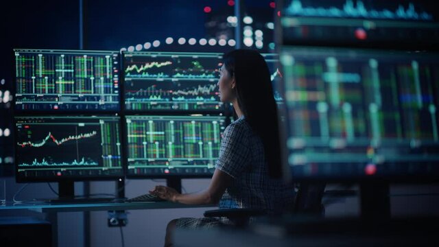 Financial Analyst Working on a Computer with Multi-Monitor Workstation with Real-Time Stocks, Commodities and Exchange Market Charts. Businesswoman at Work in Investment Broker Agency Office at Night.