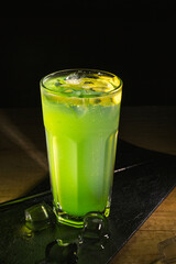 Glass of green refreshing cold kiwi cocktail served with ice cubes on slate board on table in dark bar at night 