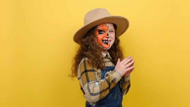 Scary bright little girl child with Halloween makeup mask wears brown hat and shirt looking camera nodding, posing isolated over yellow color background in studio. Celebration holiday party concept