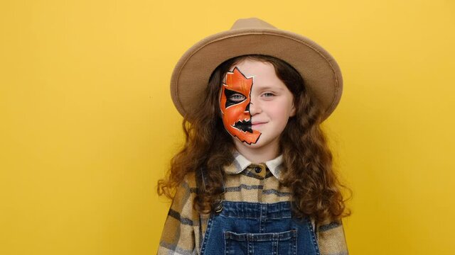 Close up portrait of bright little girl child with Halloween makeup mask wears hat and shirt, keep eyes closed open smile isolated on yellow color background in studio. Celebration party concept