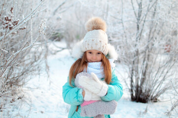 Fototapeta premium A small, charming girl in winter in a snowy park holds a teddy bear. Background of a winter forest landscape