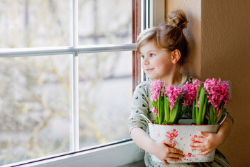 Little toddler girl sitting by window with blossoming pink hyacinth flowers. Happy child, indoors. Mother's day, valentine's day or birthday and spring concept. © Irina Schmidt