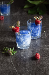 Shot with an alcoholic cocktail of bright blue color with a raspberry decoration on a gray concrete background