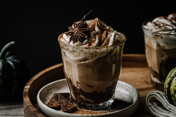 Two glasses of Pumpkin pie spice mocha latte with whipped cream and dark chocolate on plate on wooden tray with pumpkins on dark background .