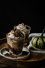 Two glasses of Pumpkin pie spice mocha latte with whipped cream and dark chocolate on plate on wooden tray with pumpkins on dark background .