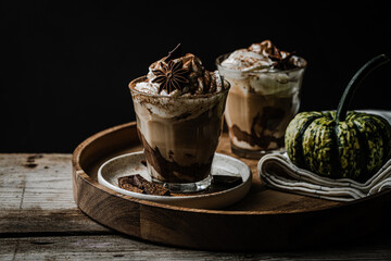Two glasses of Pumpkin pie spice mocha latte with whipped cream and dark chocolate on plate on wooden tray with pumpkins on dark background .