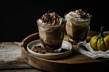 Two glasses of Pumpkin pie spice mocha latte with whipped cream and dark chocolate on plate on wooden tray with pumpkins on dark background .