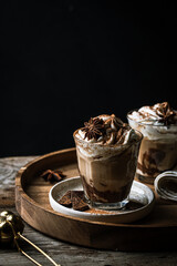 Two glasses of Pumpkin pie spice mocha latte with whipped cream and dark chocolate on plate on wooden tray on dark background .