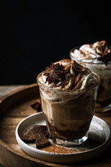 Two glasses of Pumpkin pie spice mocha latte with whipped cream and dark chocolate on plate on wooden tray on dark background .