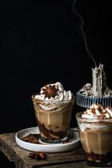 Pumpkin pie spice mocha latte with whipped cream in glass, dark chocolate on plate and natural wax candle on wooden table on dark background .
