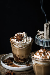 Pumpkin pie spice mocha latte with whipped cream in glass, dark chocolate on plate and natural wax candle on wooden table on dark background .