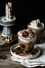 Two glasses of Pumpkin pie spice mocha latte with whipped cream, dark chocolate on plate and natural wax candle on wooden table on dark background .