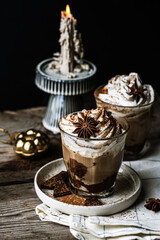 Two glasses of Pumpkin pie spice mocha latte with whipped cream, dark chocolate on plate and natural wax candle on wooden table on dark background .