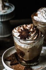 Two glasses of Pumpkin pie spice mocha latte with whipped cream, dark chocolate on plate and natural wax candle on wooden table on dark background .