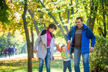 Obraz premium Beautiful young family on a walk in autumn forest on maple yellow trees background. Father and mother hold son on hands. Happy family leisure together concept