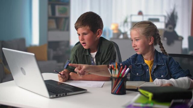 children learn online, cute male pupil studying with girl with plaster cast on the hand while sitting at home at the table with computer laptop