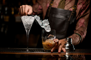 beautiful clear martini glass full of ice and glass of shaker with cocktail in which pouring ice cubes are on bar counter