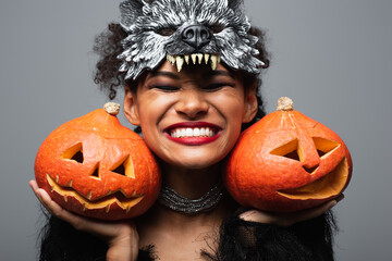 overjoyed african american woman in wolf halloween mask holding carved pumpkins and smiling with...