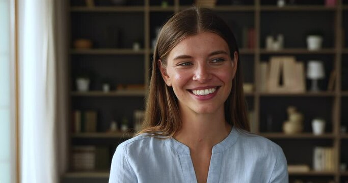 Head shot portrait of cheerful pretty young adult gen z woman standing at home indoors looking at camera. Happy optimistic satisfied teen female posing for webcam with shiny white toothy smile on face