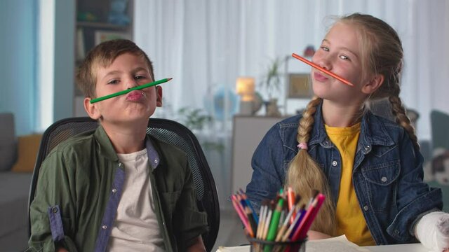 cheerful children with colored pencils have fun together, close up happy boy and girl fool around during learning at home sitting at table