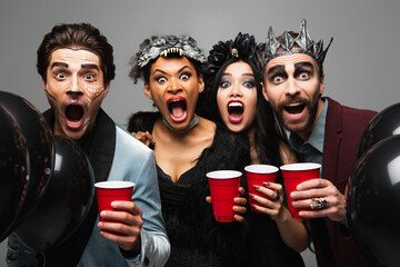 spooky multiethnic friends with plastic cups screaming and growling at camera on halloween party...