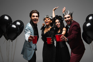 excited multiethnic friends in halloween costumes holding plastic cups and waving hands isolated on grey