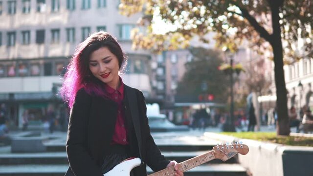Daring rebel woman with purple hair playing electric guitar at city center during bright sunset. People walking in background. Asian girl like rock star performing show at the street. Music lifestyle