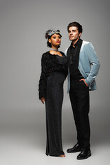 confident and elegant interracial couple in halloween costumes looking at camera on grey