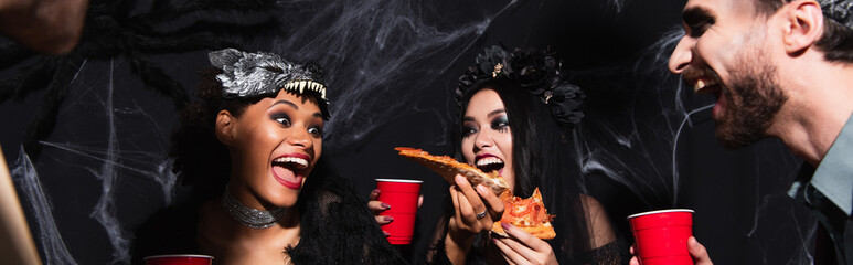 thrilled african ameican woman holding pizza during halloween party with multiethnic friends on...